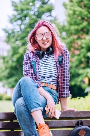 Beautiful modern young female teenager Portrait with extraordinary hairstyle color in a checkered shirt with wireless headphones on the neck sitting on the city park bench and smiling at the camera.の写真素材