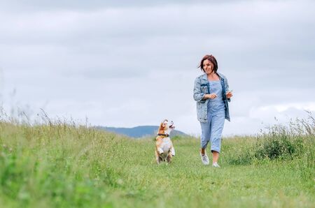 Happy smiling jogging female with fluttering hairs and her beagle dog running and looking at eyes. Walking by meadow grass path in nature with pets, healthy active people lifestyle concept image.の写真素材