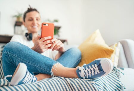 Young female sitting on cozy comfortable living room sofa, smiling and chatting with friends using modern smartphone. Selective focus on new blue sneakers. Comfortable clothing and footwear conceptの写真素材
