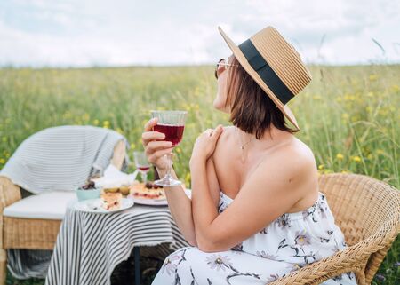 Young Woman in sunglasses and  straw hat dressed light summer dress sitting in rattan chair on the high green grass meadow near picnic table cheerful, enjoying wine and landscape.Natural beauty imageの写真素材