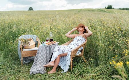 Young Woman dressed light summer dress and straw hat sitting in a rattan chair on the high green grass meadow and enjoying the moment near the picnic table with red wine and homemade pie.の写真素材