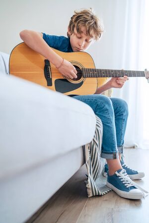 Preteen boy playing acoustic guitar dressed casual jeans, shirt and new sneakers sitting on the cozy sofa at home living room. Music education top angle view concept vertical image.の写真素材