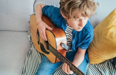 Preteen boy playing acoustic guitar dressed casual jeans, blue shirt sitting on the cozy sofa at the home living room and thinking about something. Music education concept Top angle view image.の写真素材
