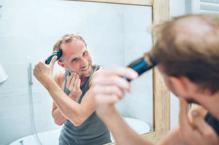 Body and skincare treatment concept. Smiling Man making new style haircut trimming a hairs using an electric rechargeable Trimmer looking in bathroom mirror. Hairstyleの写真素材