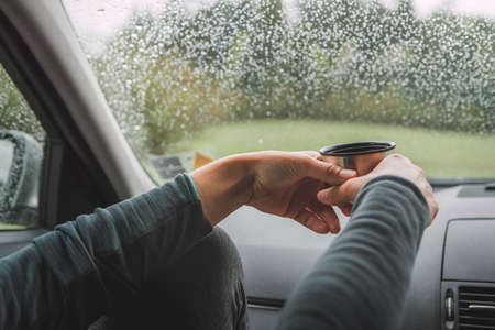 Close up shot of graceful female palms holding the hot tea tourist mug. She sitting on co-driver seat inside modern car, enjoying the moody rainy day weather looking through windscreen drops.の写真素材