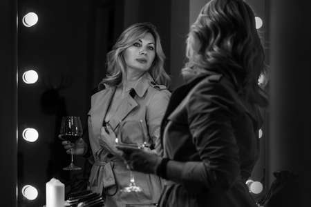 Sensual portrait of middle aged seductive woman in Trench coat gazing at mirror reflection appreciating her hair style. Human emotion or clothing fashion concept image. Back view selective focus shot.の写真素材