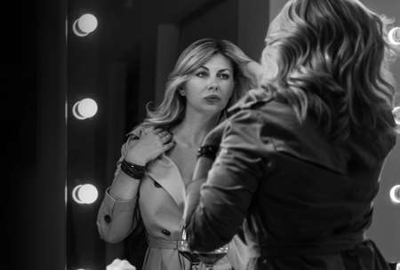 Sensual portrait of middle aged seductive woman in Trench coat gazing at mirror reflection appreciating her hair style. Human emotion or clothing fashion concept image. Back view selective focus shot.の写真素材