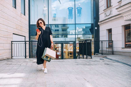 Smiling beautiful modern middle-aged female Portrait dressed black dress and sunglasses with shopping bags browsing slim smartphone listening music with wired earphones. Beautiful people concept imageの写真素材