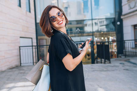 Beautiful modern middle-aged female Smiling Portrait dressed black dress and sunglasses with shopping bags holding slim smartphone listening music with wired earphones. Beautiful people concept image.の写真素材