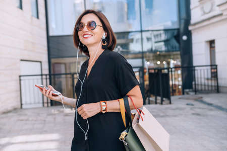 Smiling beautiful modern middle-aged female Portrait dressed black dress and sunglasses with shopping bags holding slim smartphone listening music with wired earphones. Beautiful people concept image.の写真素材