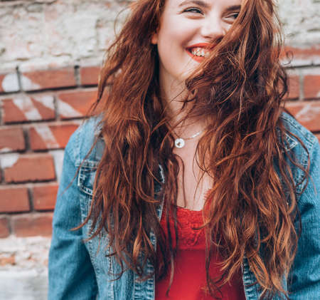 Sincerely smiling Red curled hair caucasian teen girl with applied red lipstick lips with blue eyes fashion vertical portrait on the red brick wall background. Natural people beauty concept image.の写真素材