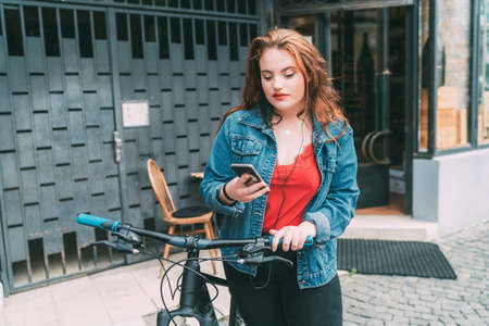 Portrait of red curled long hair caucasian teen girl on the city street walking with bicycle using the smartphone with earphones. Natural people beauty urban life concept image.の写真素材