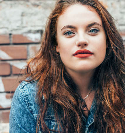 Red curled hair caucasian teen girl with applied red lipstick lips with blue eyes fashion portrait on the red brick wall background. Natural people beauty concept image.の写真素材