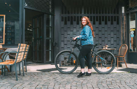 Fashion portrait of red curled long hair caucasian teen girl walking with bicycle on the city street. Natural people beauty urban life concept image.の写真素材