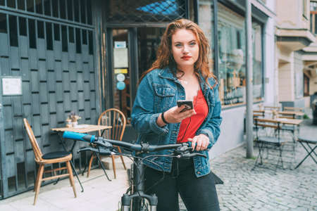 Red curled long hair caucasian teen girl on the city street walking with bicycle using the smartphone with earphones fashion portrait. Natural people beauty urban life concept image.の写真素材