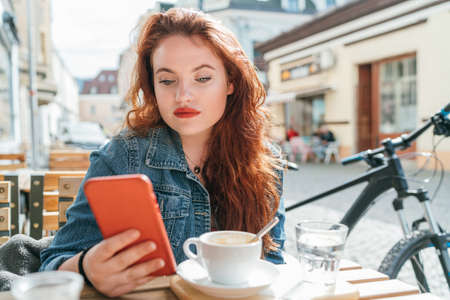 Portrait of red curled long hair caucasian teen girl sitting on a cozy cafe outdoor terrace on the street using the modern smartphone. Young woman taking a break in her city bicycle tour.の写真素材