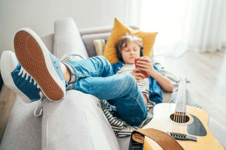 Preteen boy lying with guitar on cozy sofa dressed casual jeans and new sneakers listening to music and chatting using wireless headphones connected with a smartphone. Using electronic devices conceptの写真素材