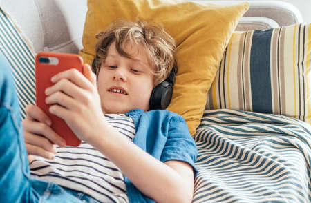 Preteen boy lying at home on cozy sofa dressed casual jeans and new sneakers listening to music and chatting using wireless headphones connected with smartphone. Child use electronic devices conceptの写真素材