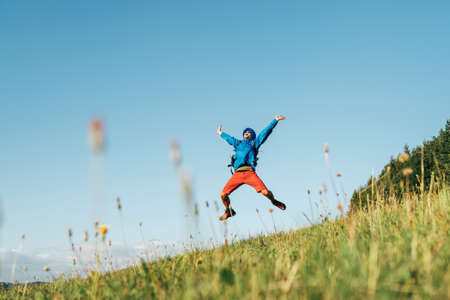 Backpacker traveler emotionally jumping over green grass mountain meadow with backpack with wide opened arms and legs. Human's freedom in nature concept image.の写真素材