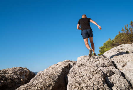 Active mountain skyrunner dressed black t-shirt and running shoes jogging the cliff summit during the morning scamper. Sporty people activities a wide-angle concept back view image.の写真素材