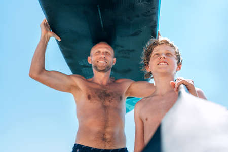 Blonde teenage son with father surfers smiling carrying the stand-up paddleboard with a paddle on the bright sunny day noon. Active family summer vacation time near the sea concept image.の写真素材