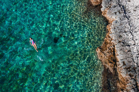 Top aerial view of the young female floating on the back and relaxing on the warm turquoise Adriatic sea waves with rocky coastline. Carefree people vacation time concept image.の写真素材