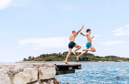 Father and son having a fun on a merry vacation days on Adriatic sea coast. They dressed swimming trunks jumping to the waives from the boat pier. Family time, childhood or fatherhood concept image.の写真素材