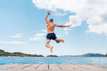 Middle-aged man dressed swimming trunks funny jumping to the waves from the boat pier as he having fun on merry vacation days on Adriatic sea coast. Carefree people vacation time concept image.の写真素材