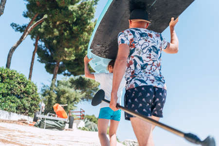 Teenage boy with father surfers carrying on the heads the stand-up paddleboard together with a paddle on the bright sunny day noon. Active family summer vacation time near the sea concept image.の写真素材
