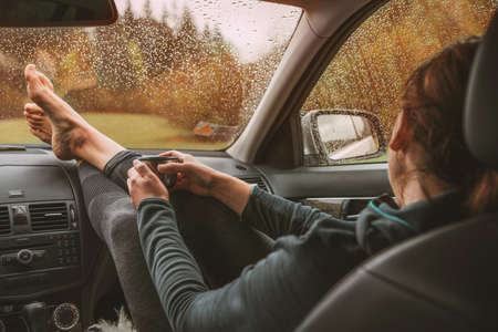 Graceful female palms holding hot tea mug. She sitting on co-driver seat inside modern car,putting legs on dashboard  enjoying a moody rainy day weather looking through windscreen drops.の写真素材