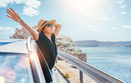 Cheerful Woman portrait enjoying the seaside road trip. Dressed a black dress, straw hat and sungllasses she wide opened arms and shining with happiness. Summer vacation traveling by auto concept.の写真素材