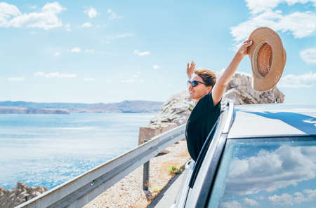 Cheerful Woman portrait enjoying the seaside road trip. Dressed a black dress with straw hat and sunglasses she wide opened arms and shining with happiness. Summer vacation traveling by auto concept.の写真素材