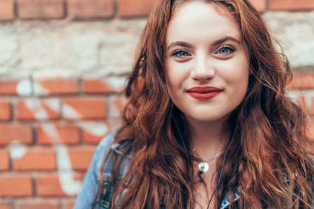 Red curled hair caucasian teen girl with applied red lipstick lips with blue eyes  smiling at camera. Fashion vertical portrait on the red brick wall background. Natural people beauty concept image.の写真素材