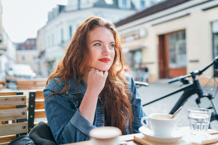 Portrait of sad red curled long hair caucasian teen girl sitting on a cozy cafe outdoor terrace on the street and looking at the street. Young woman taking a break in her city bicycle tour.の写真素材