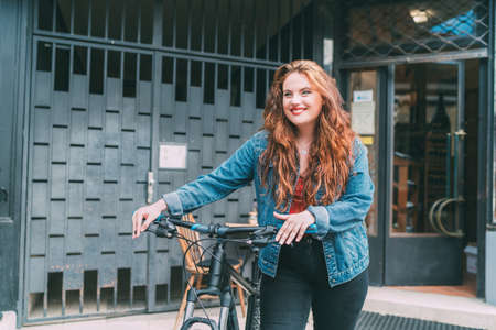 Fashion portrait of smiling Red curled long hair caucasian teen girl on the city street walking with bicycle. Natural people beauty urban life concept image.の写真素材