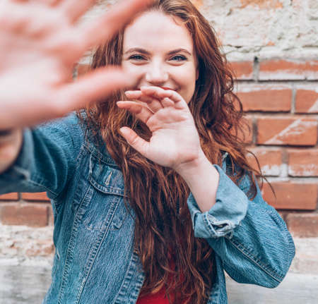 Portrait of sincerely smiling red curled long hair caucasian teen girl with applied red lipstick lips with blue eyes with a red brick wall background doing a "No photo" gesture.の写真素材