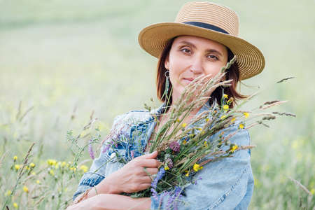 Portrait of a calm young woman with wildflowers bouquet. She dressed a jeans jacket, straw hat, and summer light dress. Natural people's beauty in the nature concept image.の写真素材