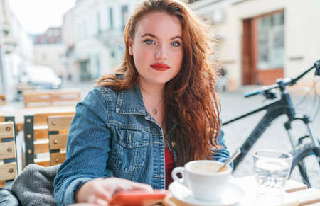 Portrait of sad red curled long hair caucasian woman sitting on a cozy cafe outdoor terrace on the street and looking at the street. Young woman taking a break in her city bicycle tour.の写真素材