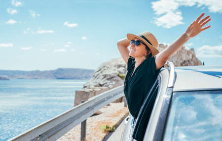 Cheerful Woman portrait enjoying the seaside road trip. Dressed a black dress, straw hat and sungllasses she wide opened arms and shining with happiness. Summer vacation traveling by auto concept.の写真素材