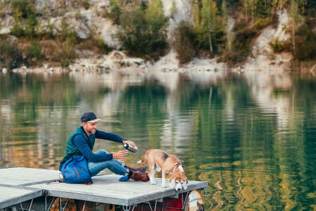 Man dog owner and friend beagle dog on the wooden pier on the mountain lake and enjoying the landscape during their walking in the autumn season time. Male pouring a tea, but dog had seen sth in waterの写真素材