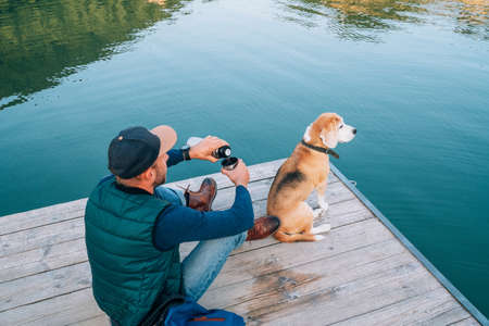 Man dog owner and friend beagle dog on the wooden pier on the mountain lake during their walking in the autumn season time. Male pouring a tea, but dog enjoying the landscape.の写真素材