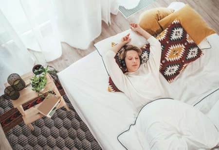 Smiling young female lying on cozy bed dressed pajamas listening to music using wireless headphones. Music playing modern technology and free time spending conceptの写真素材
