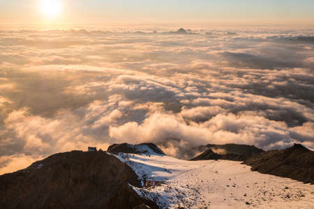 French Alps breathtaking over the clouds sunset sky landscape with a mountain hut on the lited...