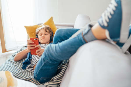 Preteen boy lying at home on cozy sofa dressed casual jeans and new sneakers listening to music and chatting using wireless headphones connected with smartphone. Child use electronic devices conceptの写真素材