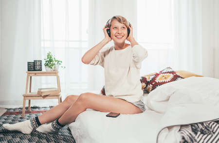 Smiling young female sitting on cozy bed dressed pajamas listening to music using wireless headphones. Music playing modern technology and free time spending conceptの写真素材