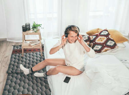 Smiling young female sitting on cozy bed dressed pajamas listening to music using wireless...