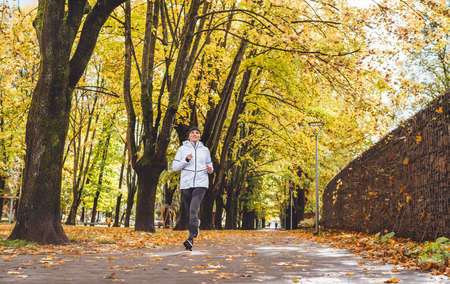Fit athletic woman have a jogging in the autumnal city park. Young fitness female smiling and running by the footway covered with yellow leaves. Active running people concept image.の写真素材