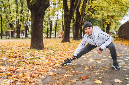 Smiling athletic woman doing workout before jogging in autumnal city park on the kids playground.Mid aged fitness female runner stretching legs while warming up. Active runners people concept imageの写真素材