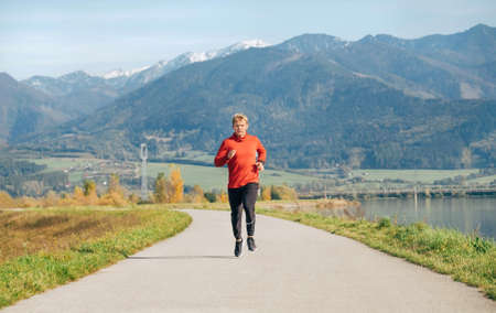 Man dressed in red long sleeve shirt, black running trousers and shoes running by the asphalt road with mountain background. Sporty people activities and a healthy lifestyle concept image.の写真素材