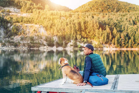 Man dog owner and his friend beagle dog are sitting on the wooden pier on the mountain lake and enjoying the landscape during their walking in the autumn season time. Human and pet concept image.の写真素材